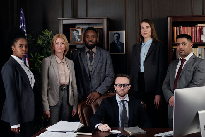 A diverse group of six professionals in formal attire stand confidently in an office setting, exuding authority and teamwork. There's an American flag and wooden bookshelves.