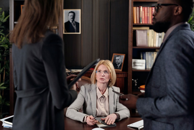 A woman seated at a desk listens attentively to two standing colleagues in an office. The setting is formal, with bookshelves and framed photos in the background.